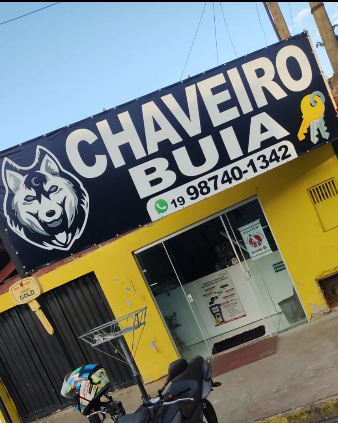 Fachada da Buia Chaveiro: placa preta com o lobo e o telefone, sobre a parede amarela da loja